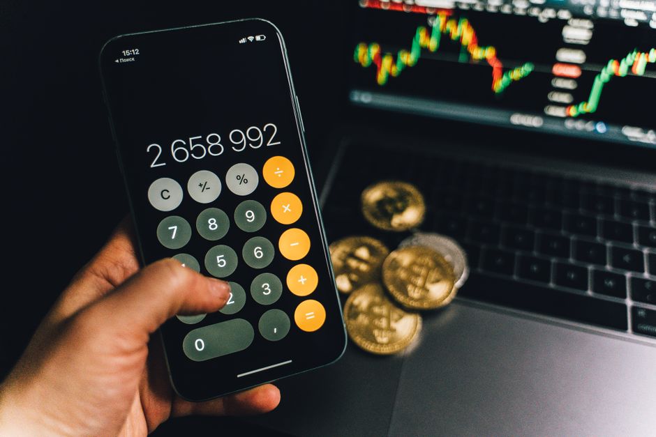 A smartphone displays a calculator app beside bitcoins and a laptop with a financial chart.