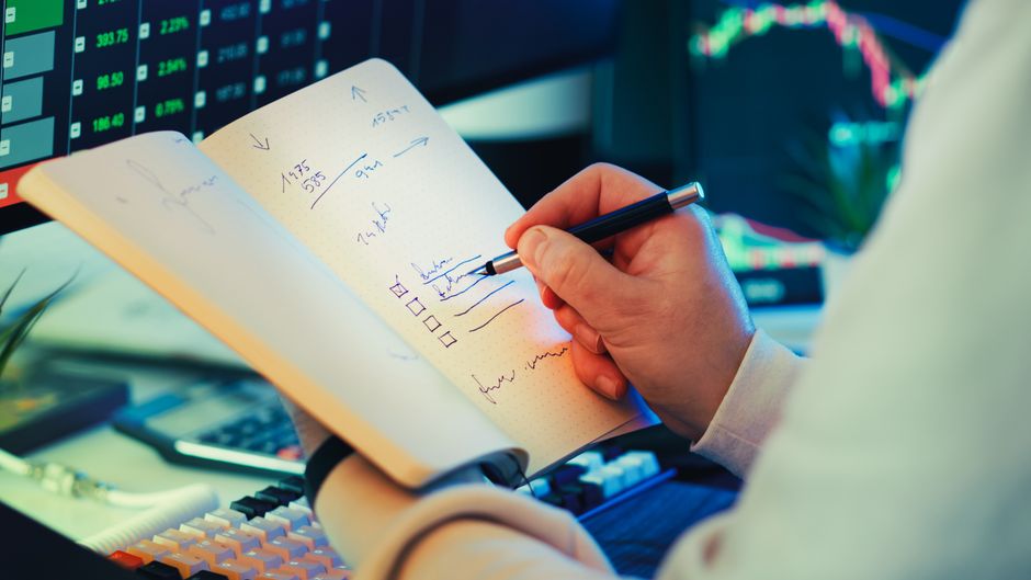 A businessman writes notes in a notebook while analyzing stock market data on a computer screen.